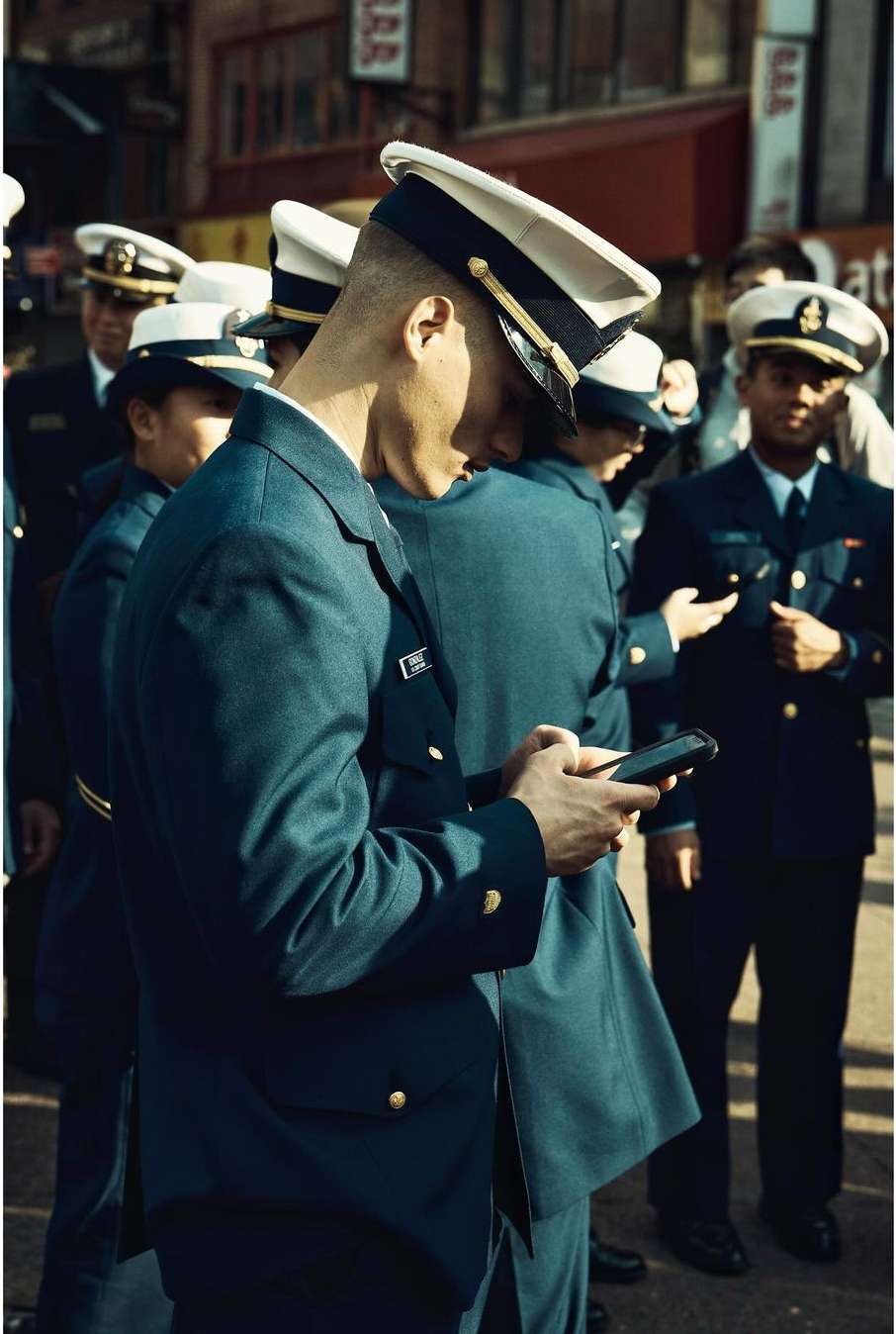 Men in uniform stand on a city street; one man uses a smartphone, and some wear white hats. Focus on the foreground subject.