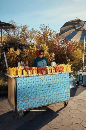 Vendor standing behind a fruit cart filled with cups of sliced mango and watermelon, with a striped umbrella overhead.