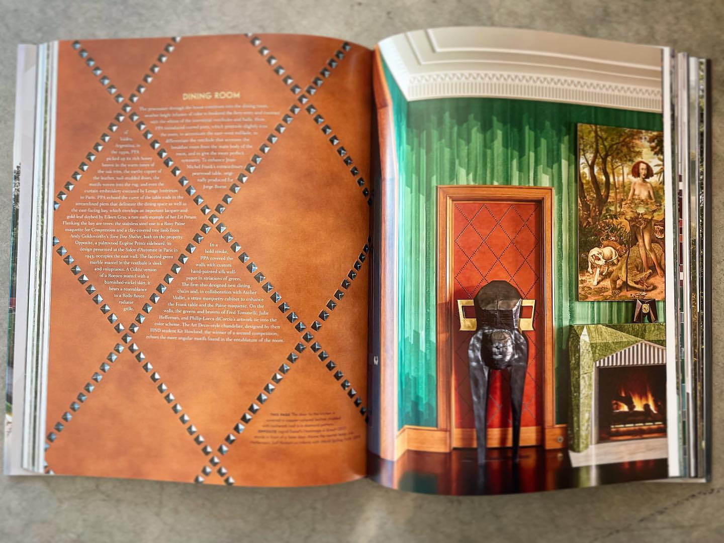 Open magazine shows an interior design spread with a paneled dining room and fireplace next to furniture and hanging art.