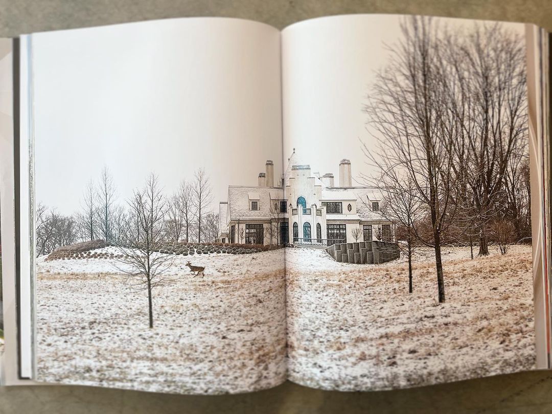 View of a large white house with many windows and chimneys on a snow-covered lawn with a deer in the foreground.