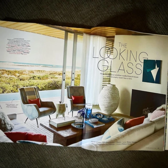 Interior design magazine spread features living room with armchairs, coffee table, sofa, view of beach, and large decorative vase.