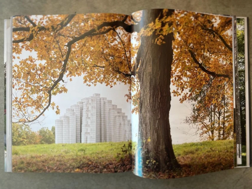 Open book featuring a photograph of a modern, blocky white monument on green grass with a large tree in autumn foliage on the right.