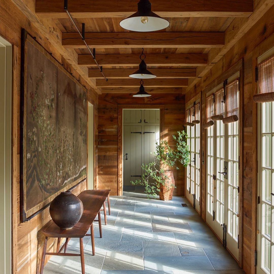 Hallway interior with light-grey stone floor, rustic wood-paneled walls and ceiling, a bench, painting, lighting fixtures, and windows.