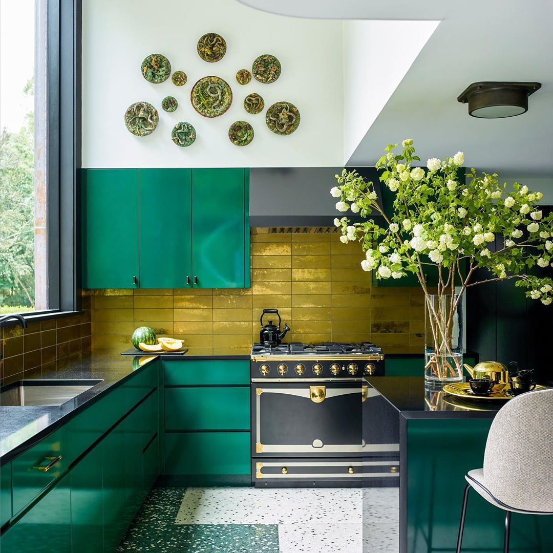 Modern kitchen interior with emerald green cabinetry, gold tile backsplash, and black appliances. Vase of white flowers on the counter.
