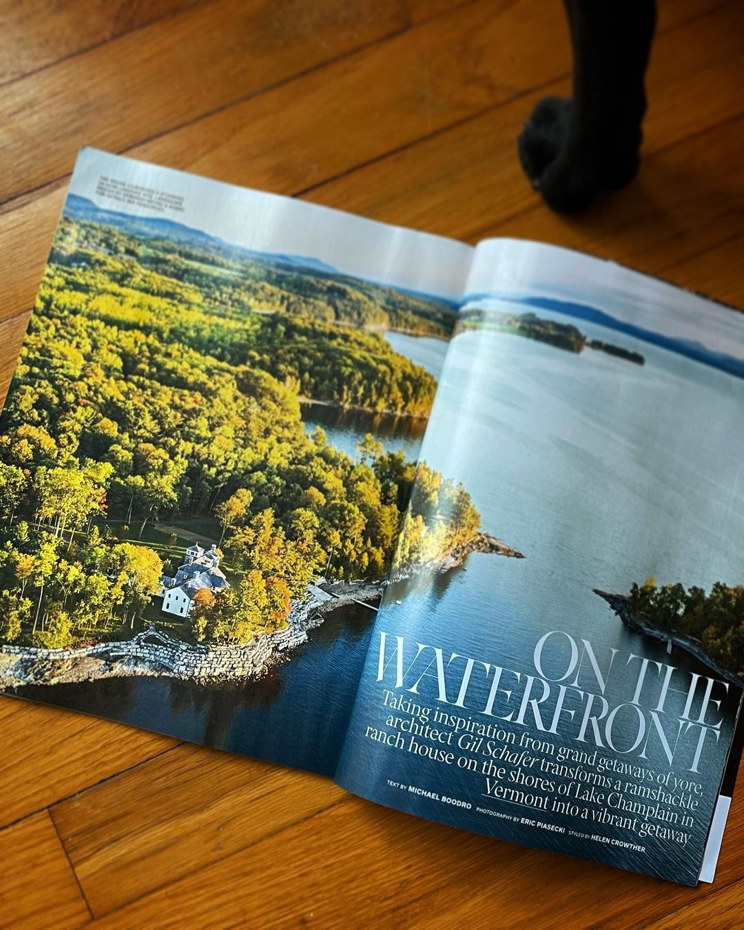 Open magazine displaying a scenic view of a lake bordered by a lush green forest and a white house, captured on a sunny day.