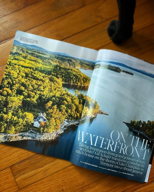 Open magazine displaying a scenic view of a lake bordered by a lush green forest and a white house, captured on a sunny day.