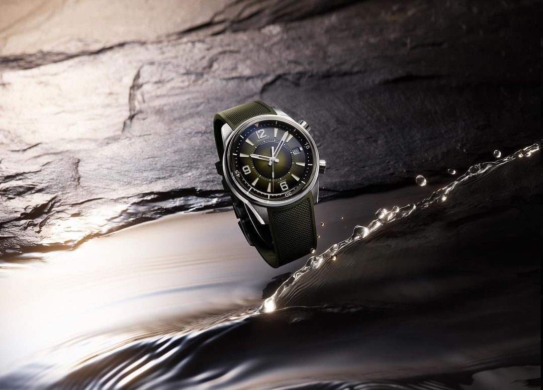 Wristwatch featuring a silver case and green strap is positioned on a water surface, set against a textured, dark stone-like backdrop.