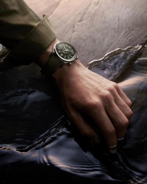 Close-up of a person's hand wearing a watch with a dark green face and matching wristband, set against a rock and water background.