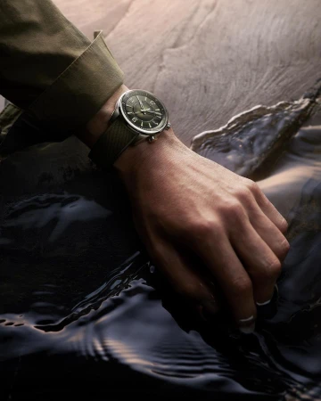 Close-up of a person's hand wearing a watch with a dark green face and matching wristband, set against a rock and water background.