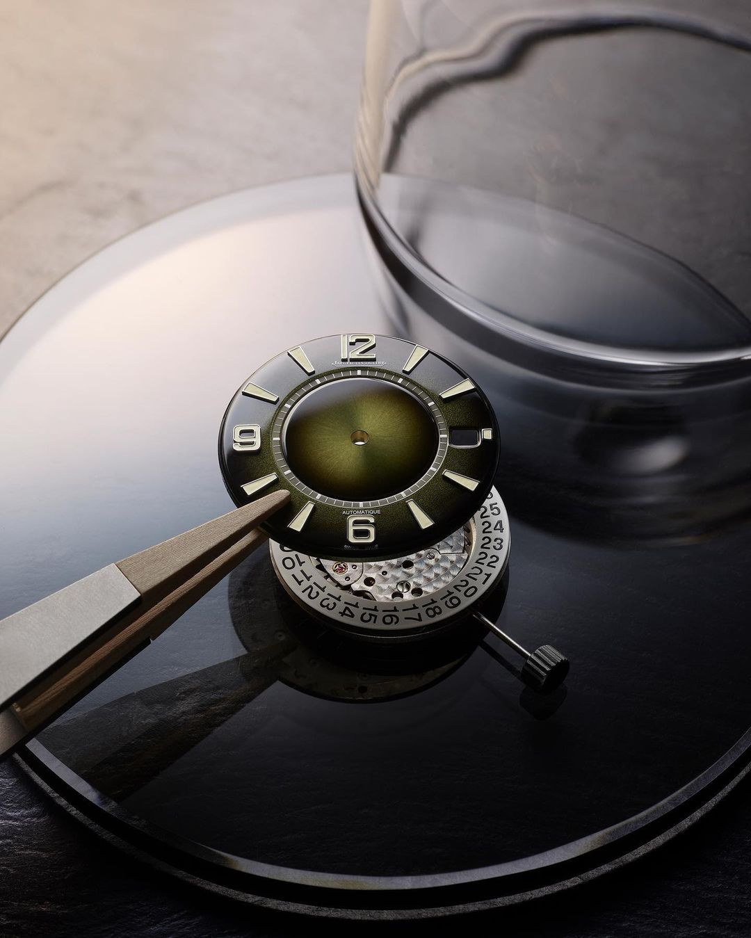 Close-up of a watch face and watch movement being assembled with tweezers on a reflective black surface with a glass cover.