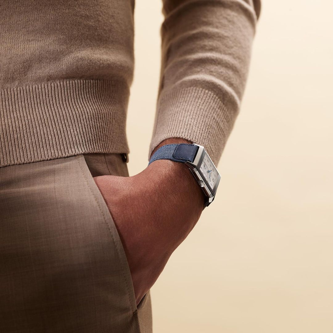 Close-up shot of a man's arm wearing a blue watch with a silver case, partially in pocket, neutral beige background, and soft lighting.