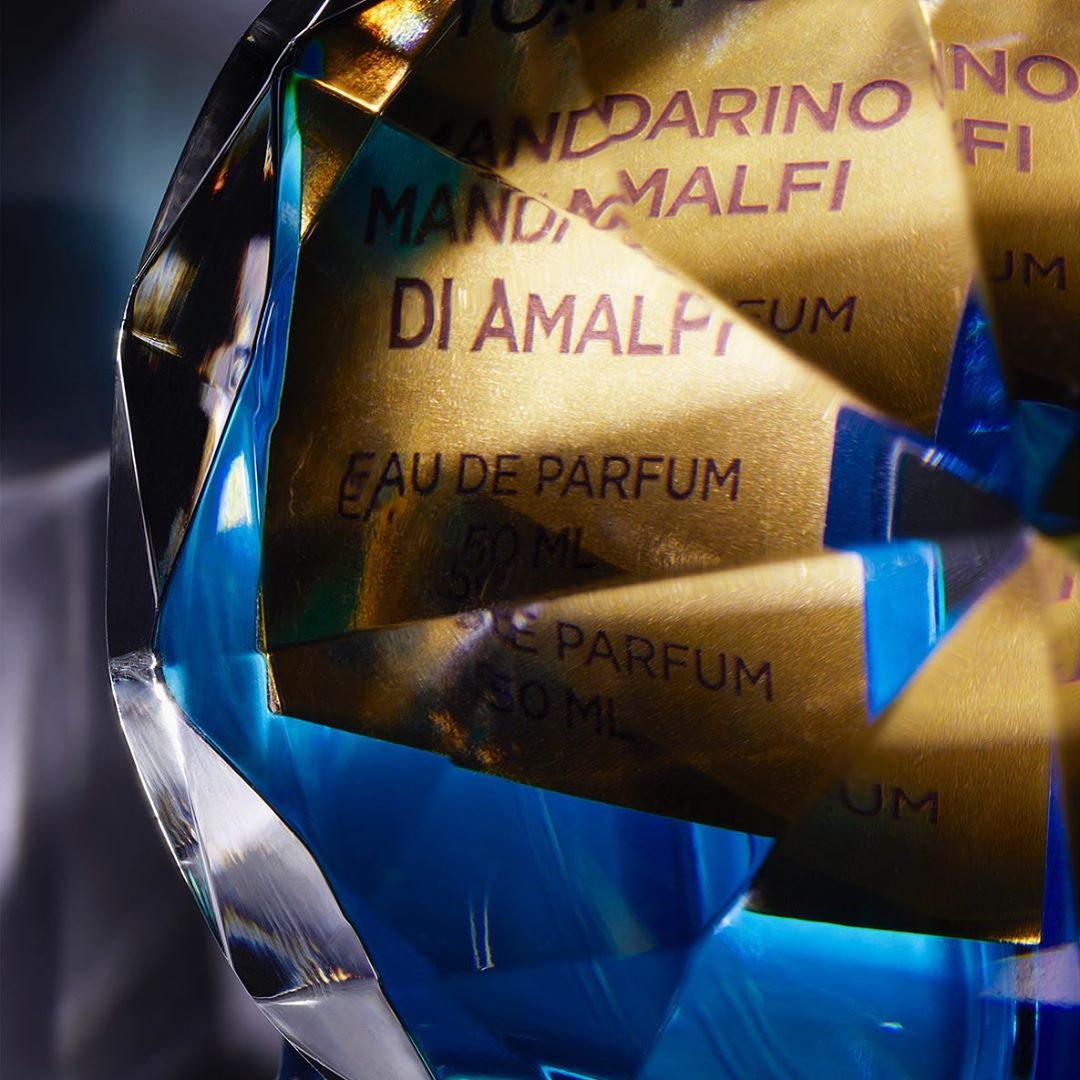 Close-up of a faceted, blue and clear perfume bottle with gold labels, featuring text 'Mandarino di Amalfi Eau de Parfum 50ml'.
