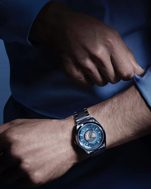 Close-up of a male wrist wearing a modern stainless steel wristwatch with blue dial, complemented by a light blue dress shirt.