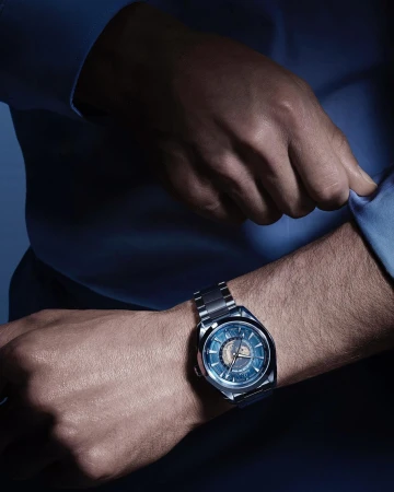 Close-up of a male wrist wearing a modern stainless steel wristwatch with blue dial, complemented by a light blue dress shirt.