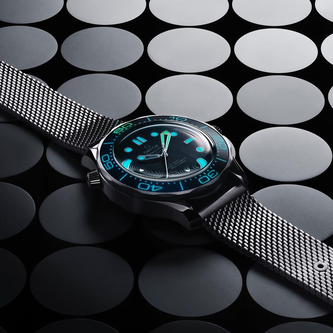 Close-up of a black and silver wristwatch with a mesh band, blue accents, set against an abstract background of dark circles.