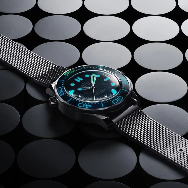 Close-up of a black and silver wristwatch with a mesh band, blue accents, set against an abstract background of dark circles.