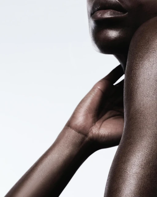 Close-up studio shot of a Black woman's shoulder, arm, and neck, showing smooth skin texture, and a neutral facial expression against white backdrop.