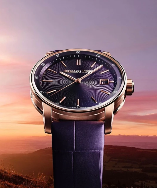 Close-up of a luxury Audemars Piguet watch with a purple face and gold casing, set against a blurred landscape backdrop.