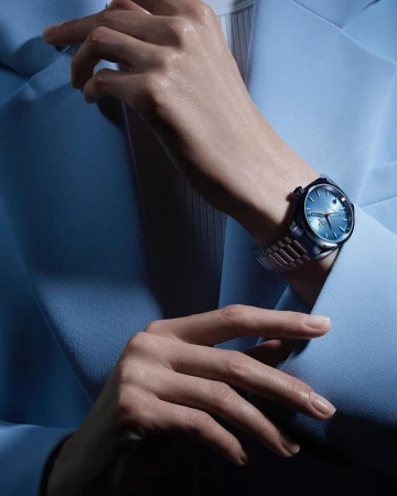 Close-up of a woman's hands wearing a blue-faced wristwatch with a silver band, set against a matching powder blue blazer.