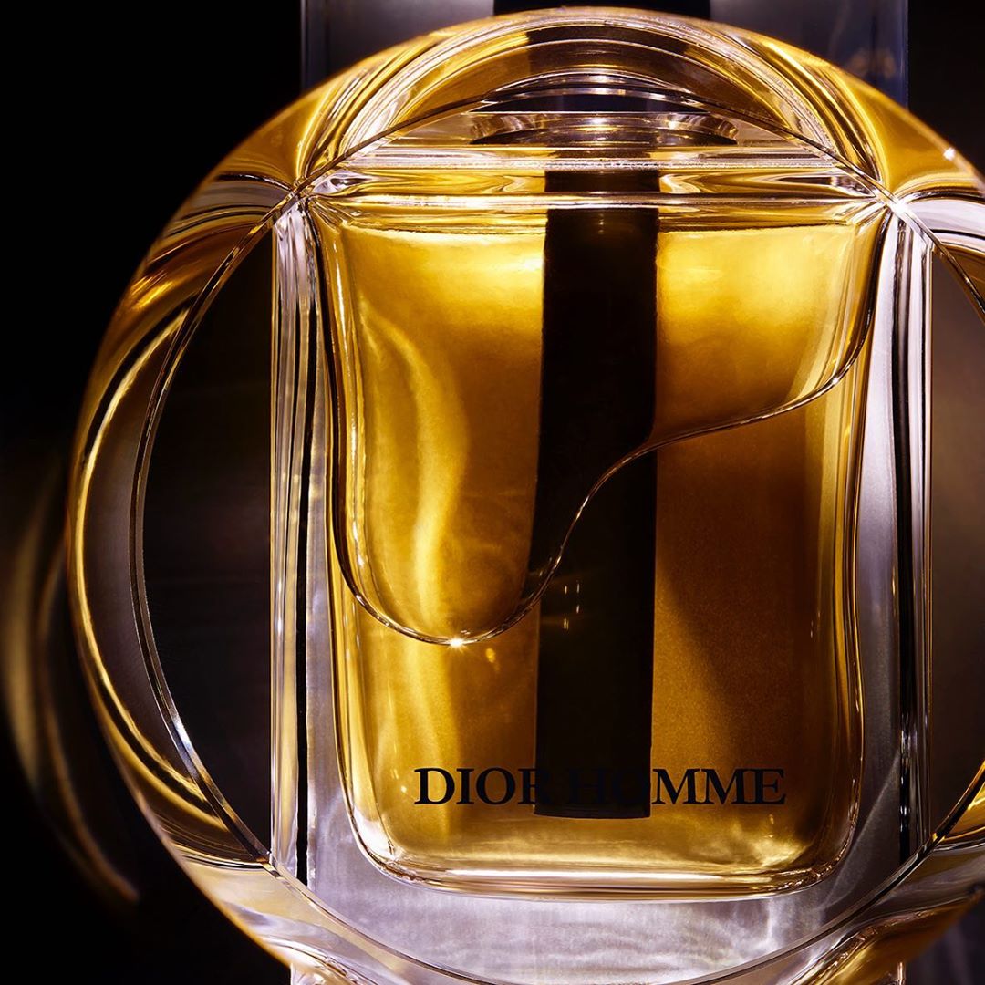Close-up of a clear, curved bottle containing golden liquid, labeled DIOR HOMME, set against a dark background. Product photography.