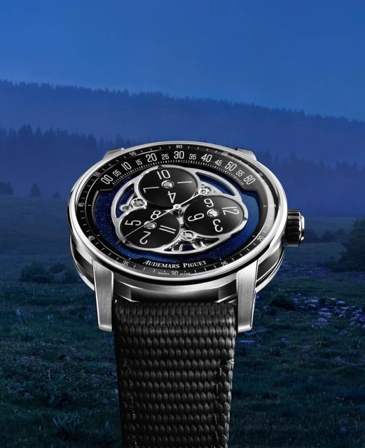 Luxury Audemars Piguet wristwatch featuring a complex dial, set against a blurred scenic backdrop of green fields and forested hills.