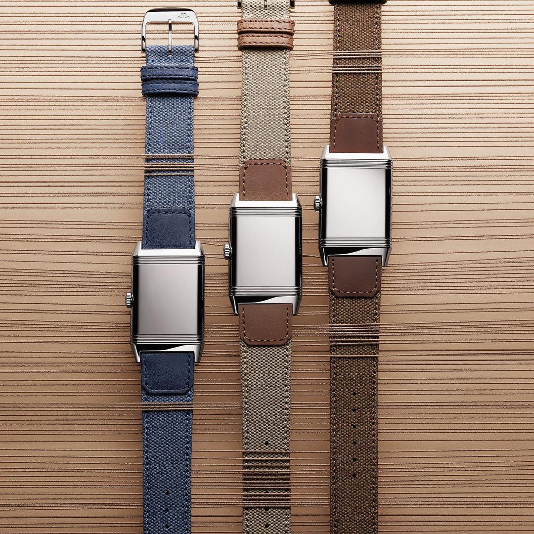 Three Jaeger-LeCoultre Reverso watches with stainless steel cases and blue, beige, and brown straps on a wood surface.