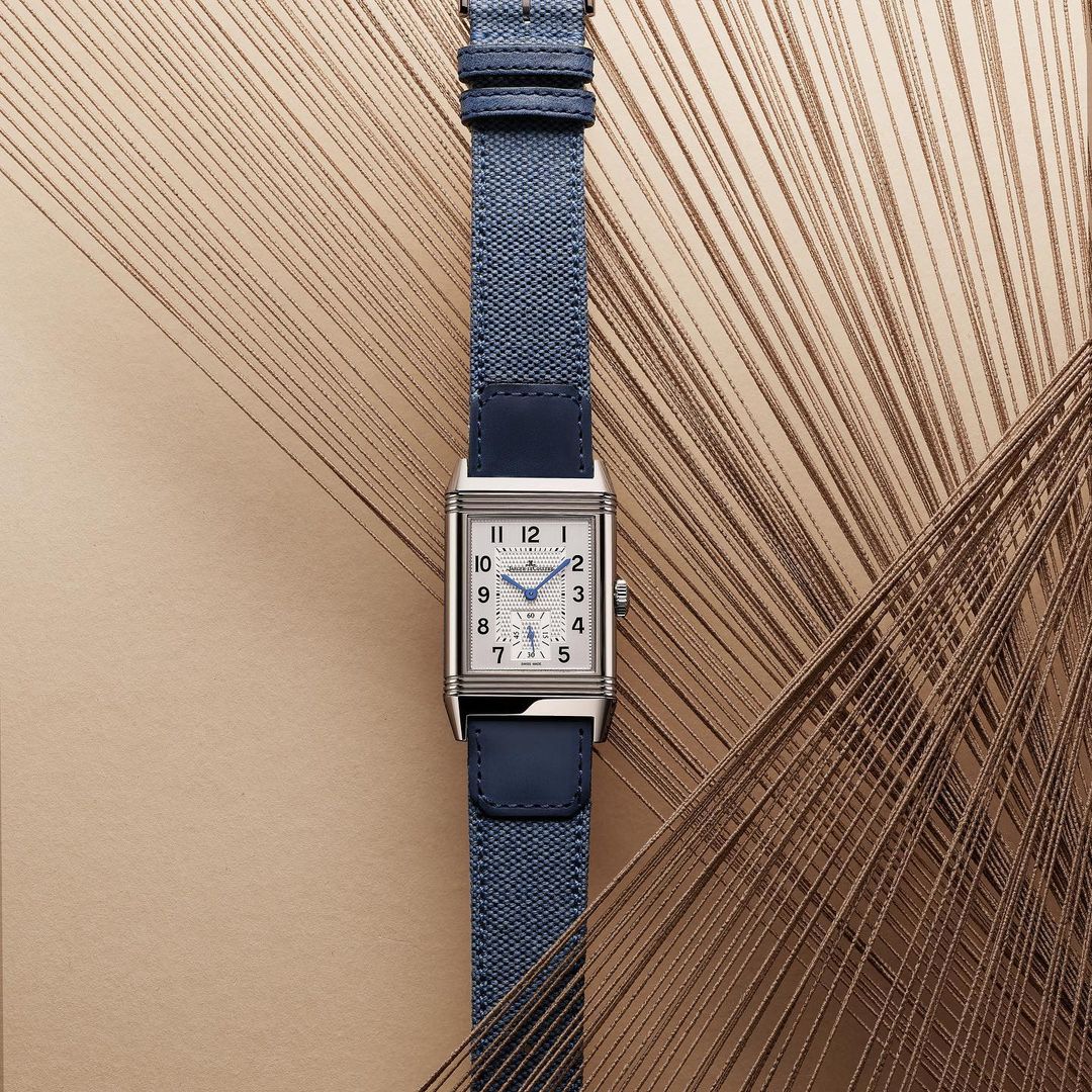 Silver Jaeger-LeCoultre Reverso Classic watch with a blue canvas strap against a neutral background with geometric thread-like accents.