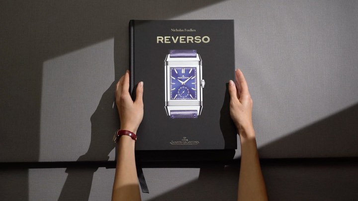 Top-down shot of hands holding a book titled 'Reverso' with a watch illustration, placed on gray fabric.