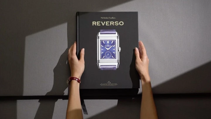Top-down shot of hands holding a book titled 'Reverso' with a watch illustration, placed on gray fabric.