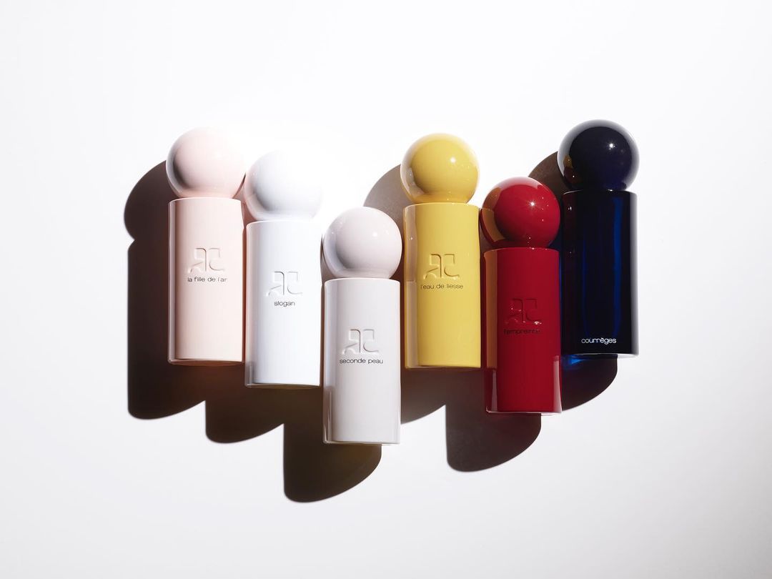 Collection of colorful Courreges perfume bottles in pink, white, yellow, red, and blue, lined up on a white surface with shadows.