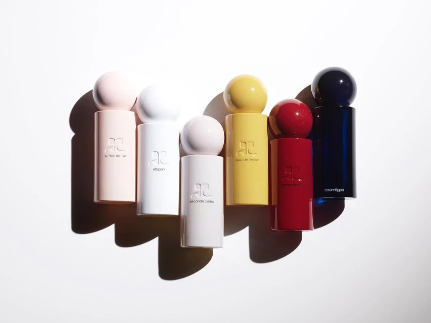 Collection of colorful Courreges perfume bottles in pink, white, yellow, red, and blue, lined up on a white surface with shadows.