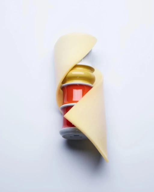 Still life shot of stacked colorful thread spools wrapped with a light yellow swatch of fabric on a plain white background.