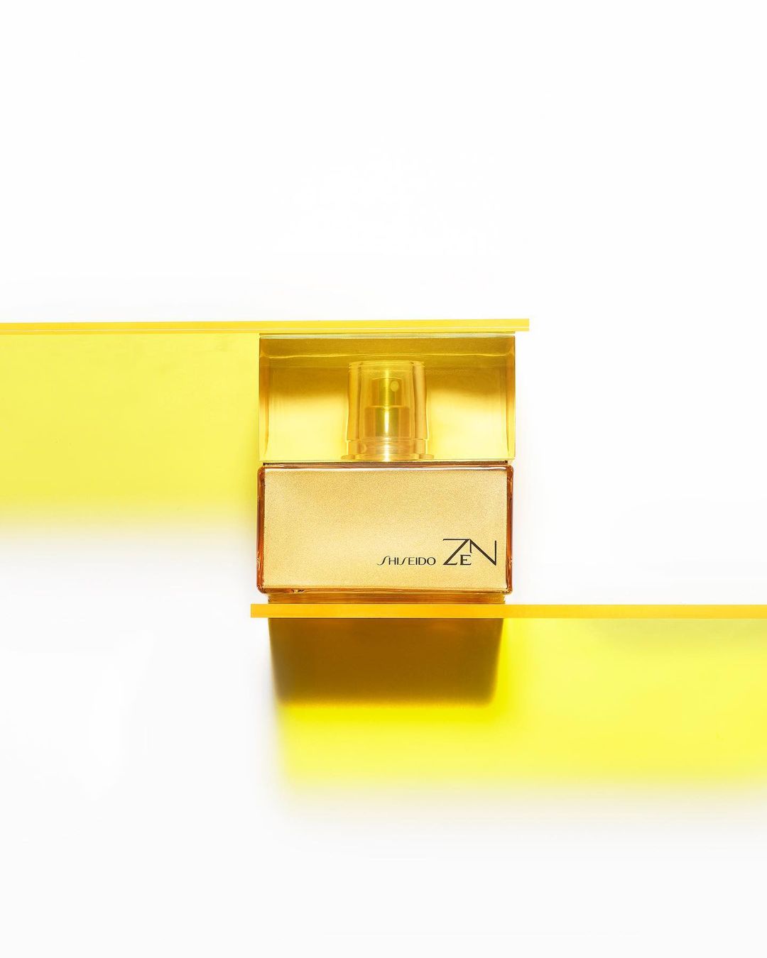 Still life shot of a gold Shiseido Zen perfume bottle on a white surface with transparent yellow bars creating a modern design.
