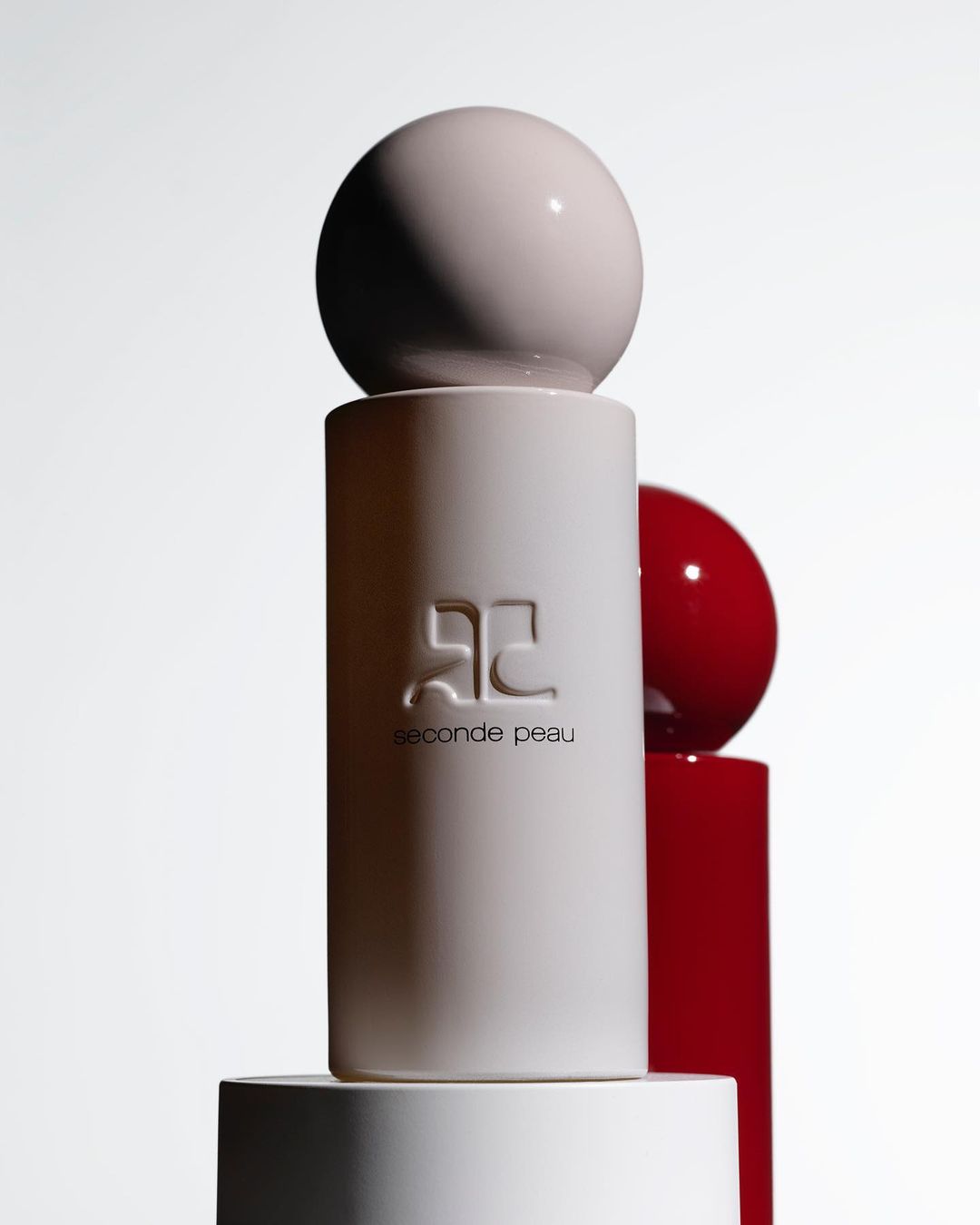 Elevated product shot of white and red spherical-topped cosmetic bottles on a layered white platform against a gradient backdrop.