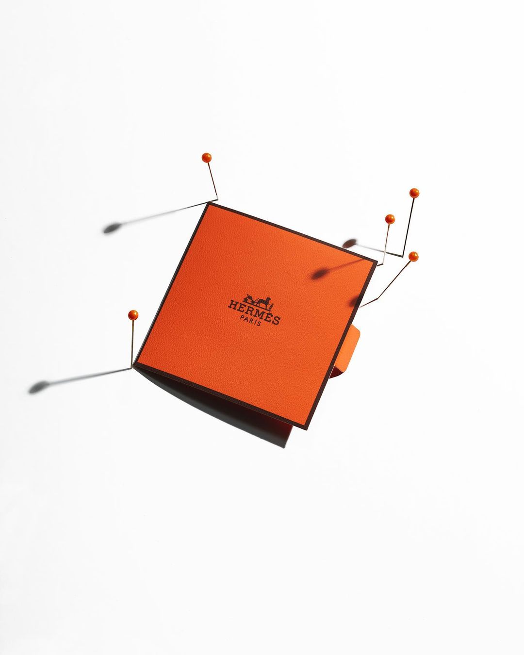 Orange Hermès box with black trim is pinned to a white surface with common orange push-pins, casting shadows, in this studio shot.