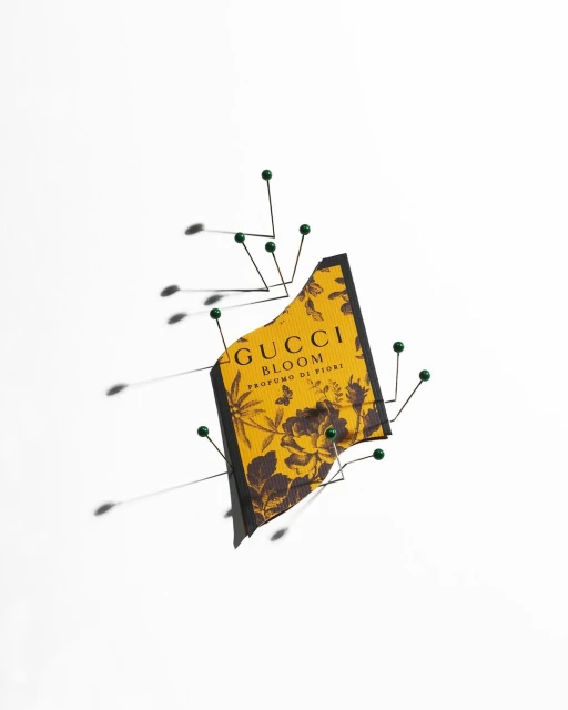 A fragrance sample from Gucci Bloom is pinned to a white surface using straight pins, with their shadows visible on the background.