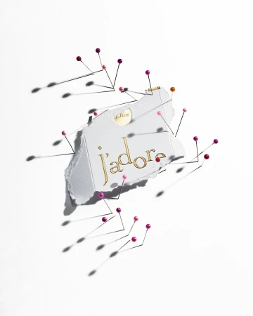 Elevated shot of Dior J'adore perfume advertisement featuring pins and white paper on a white background; beauty and product photography.