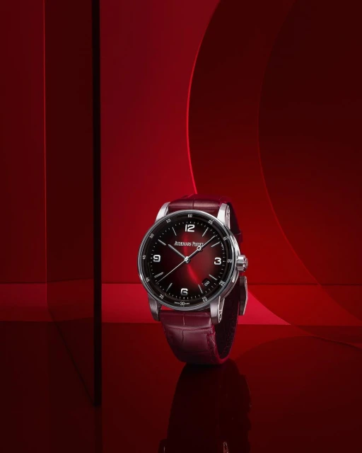 Audemars Piguet wristwatch on a reflective surface, with a red dial and a red leather strap, against a red background with geometric shapes.