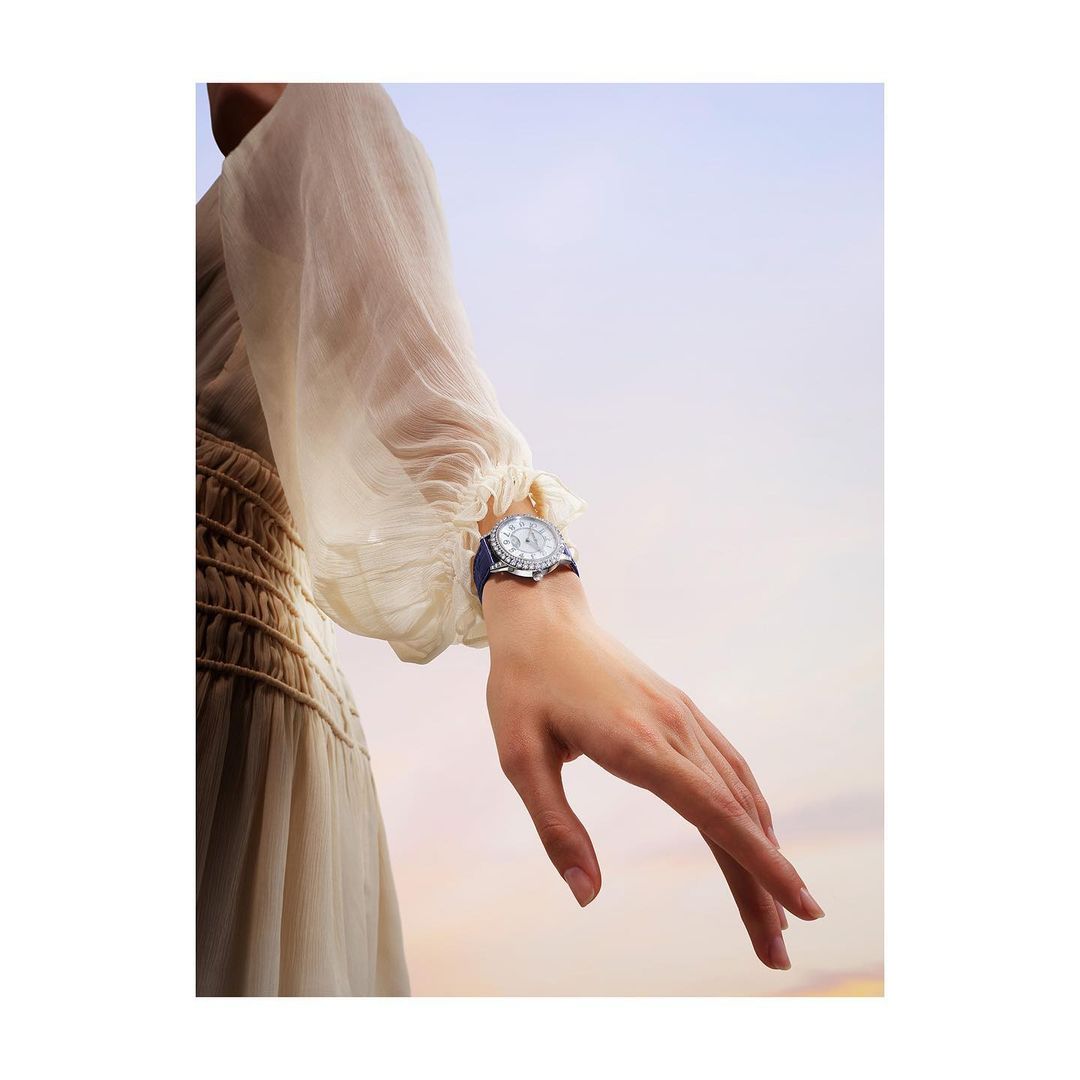 Woman's arm and wrist displaying a wristwatch with a blue strap, set against a soft gradient sky, showcasing a blend of fashion and precision.