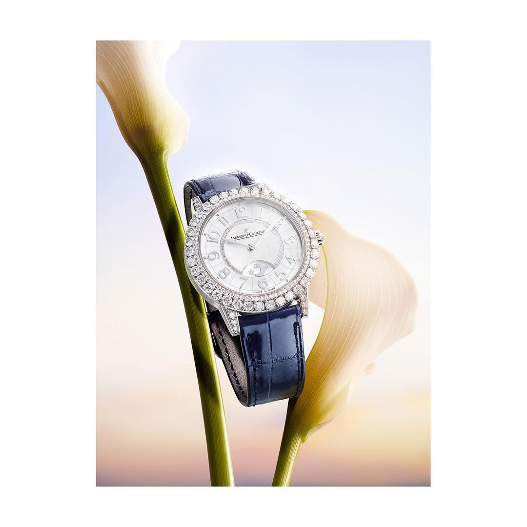 Elegant diamond-encrusted wristwatch with a blue leather strap displayed alongside calla lilies in a bright, soft-focus setting.