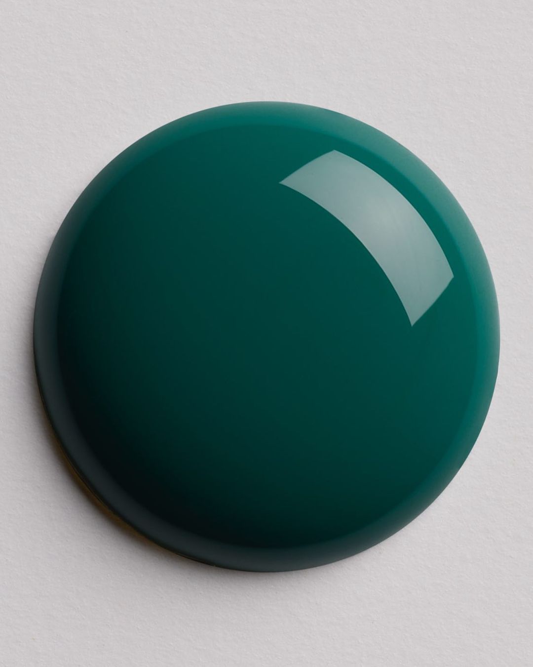 High-angle studio shot of a glossy, dark teal-colored gel nail polish droplet against a pale beige surface with subtle texture.