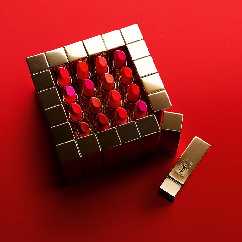 Array of YSL lipstick tubes in varying red shades arranged in gold square casing on a red backdrop, promoting cosmetics and beauty.