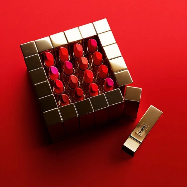 Array of YSL lipstick tubes in varying red shades arranged in gold square casing on a red backdrop, promoting cosmetics and beauty.