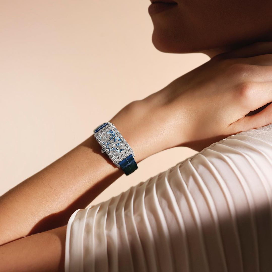 Woman's wrist adorned with a blue and silver jeweled watch, complemented by a white pleated garment against a peach background.