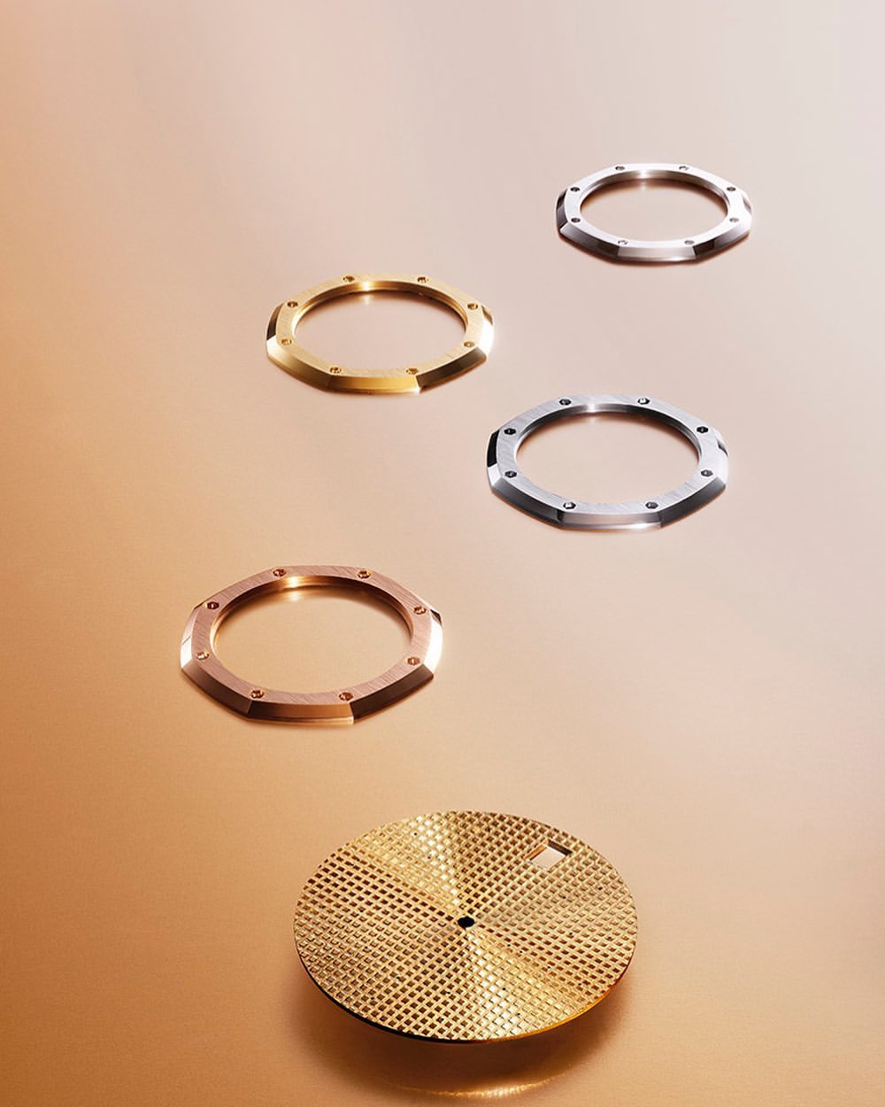 Arrangement of watch bezels and a textured watch face in gold, rose gold, and silver tones on a light brown surface, studio setting.