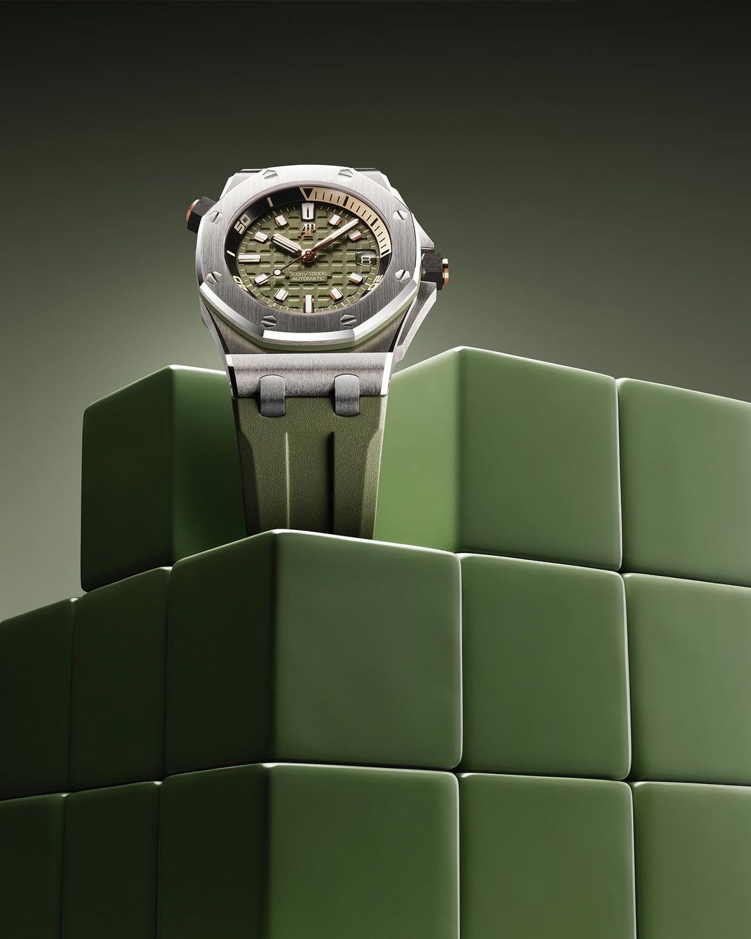 Silver wristwatch with an olive green face and band sits atop a geometric cube display against a graduated olive green background.