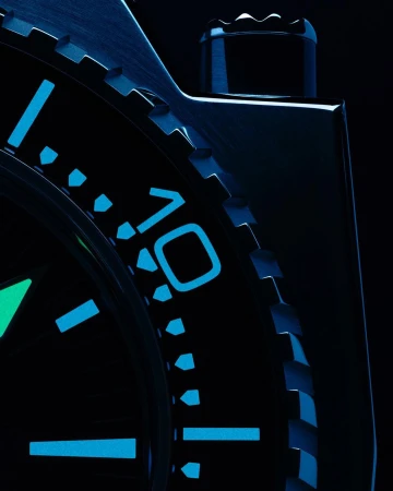 Close-up product shot of a black wristwatch featuring blue-glowing indices and numbers. Shot focuses on the watch's dial and bezel.
