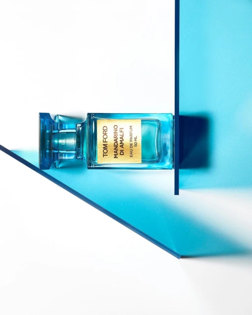 A bottle of Tom Ford Mandarino Di Amalfi eau de parfum is displayed with blue transparent geometric shapes on a white surface.