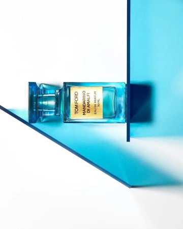 A bottle of Tom Ford Mandarino Di Amalfi eau de parfum is displayed with blue transparent geometric shapes on a white surface.