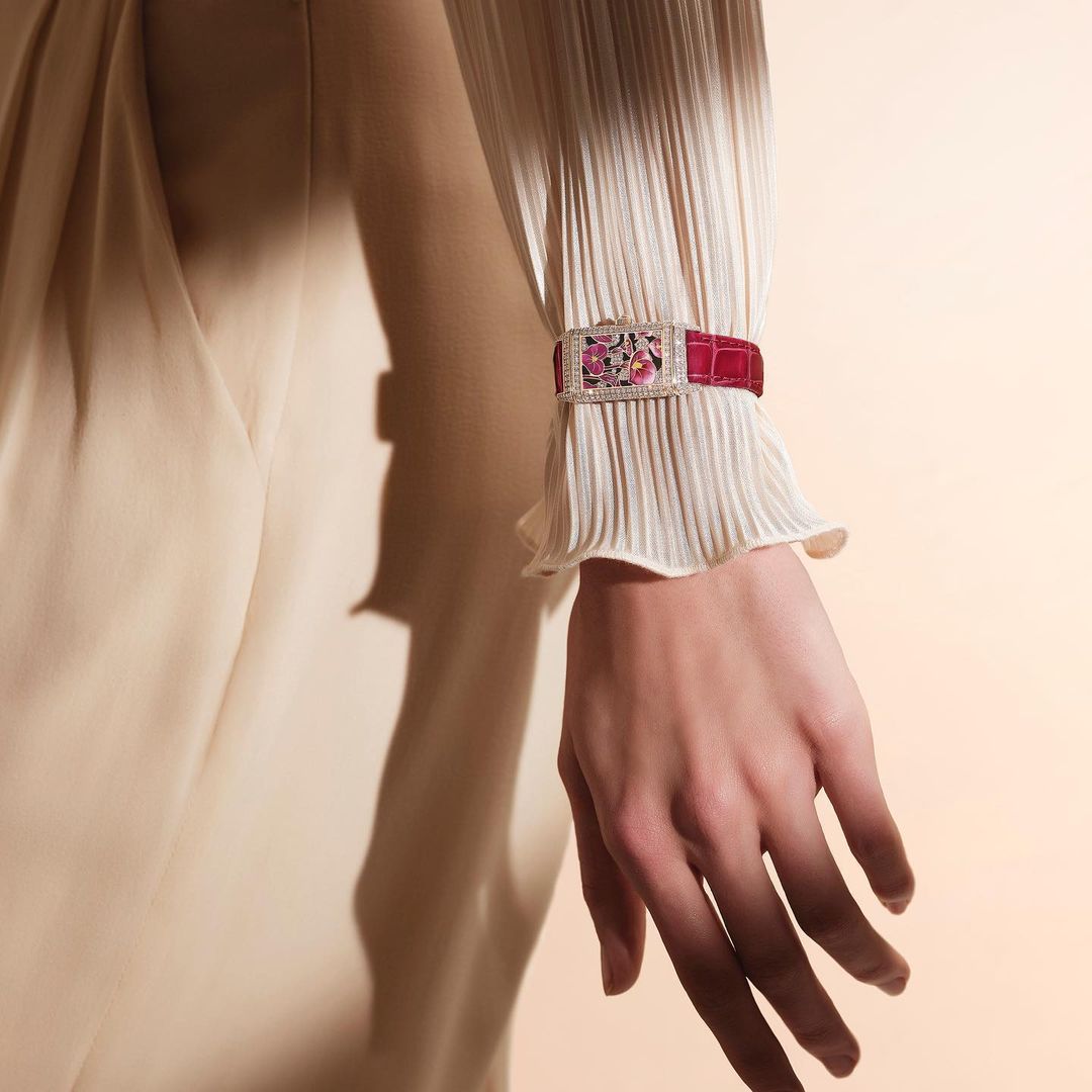Close-up of an adult female's wrist wearing a decorative wristwatch with red band, pleated sleeve, and a soft tan background. 